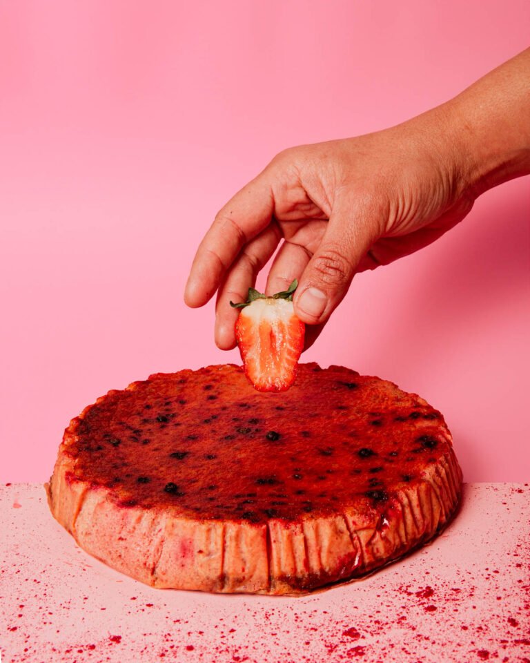 Mano colocando una fresa sobre una tarta de queso, fondo rosa